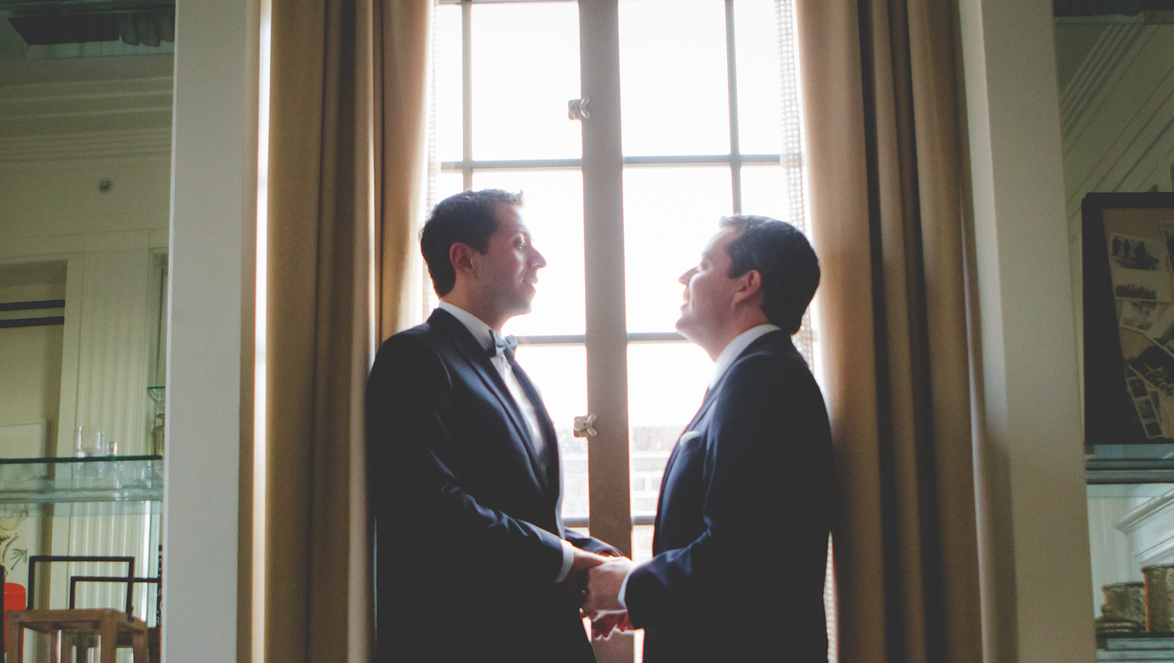 Two grooms, looking into each others eyes, stand holding hands by a window